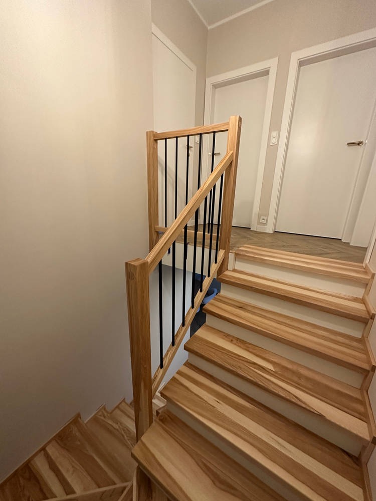 Nowoczesne schody drewniane z balustradą z jasnego drewna i czarnymi, pionowymi szczeblami, widok z góry na dół, z trzema białymi drzwiami w tle.