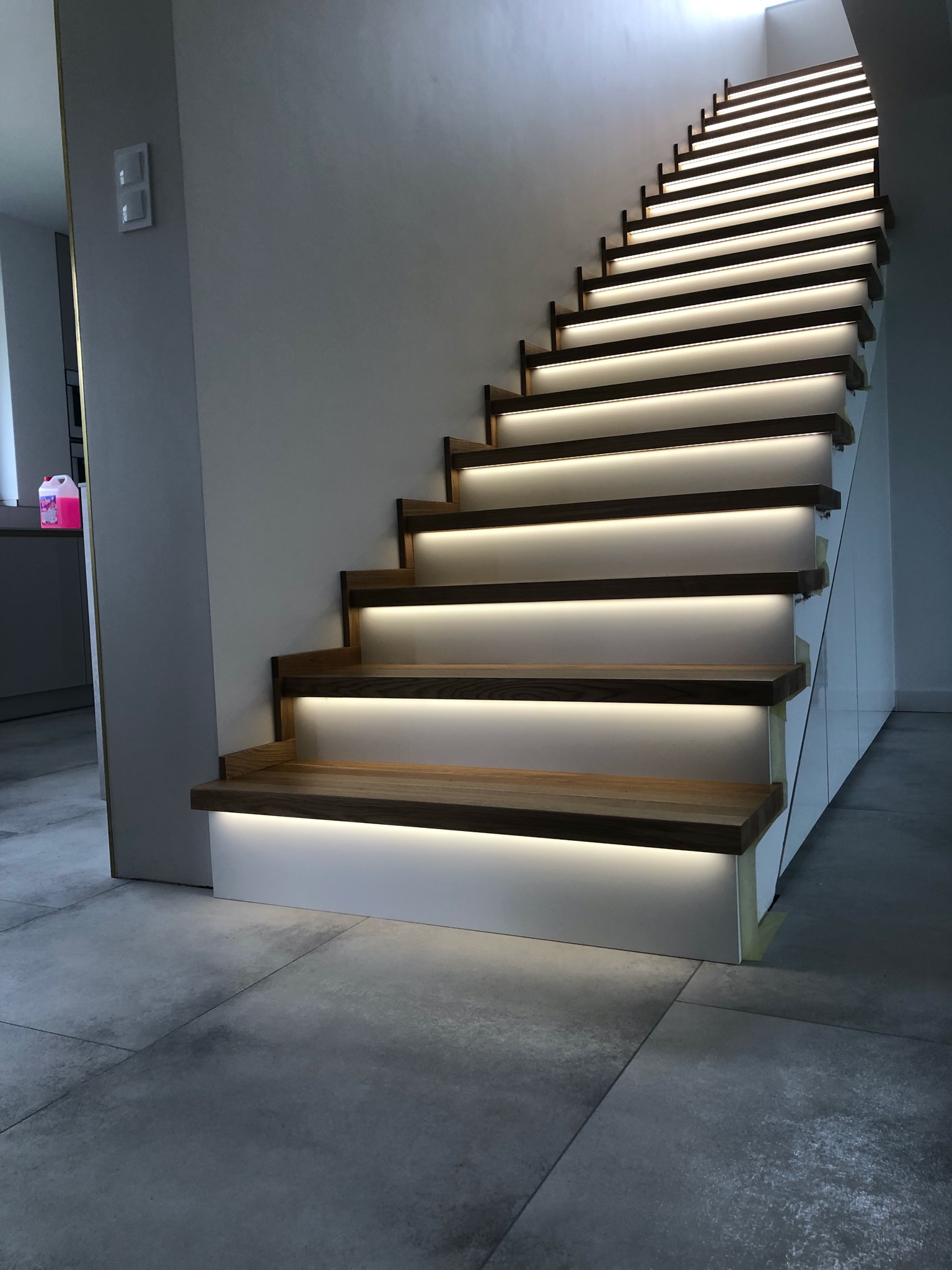 Nowoczesne schody z drewnianymi stopniami i podświetleniem LED, zamontowane przy jasnej ścianie, widok z dołu na górę.