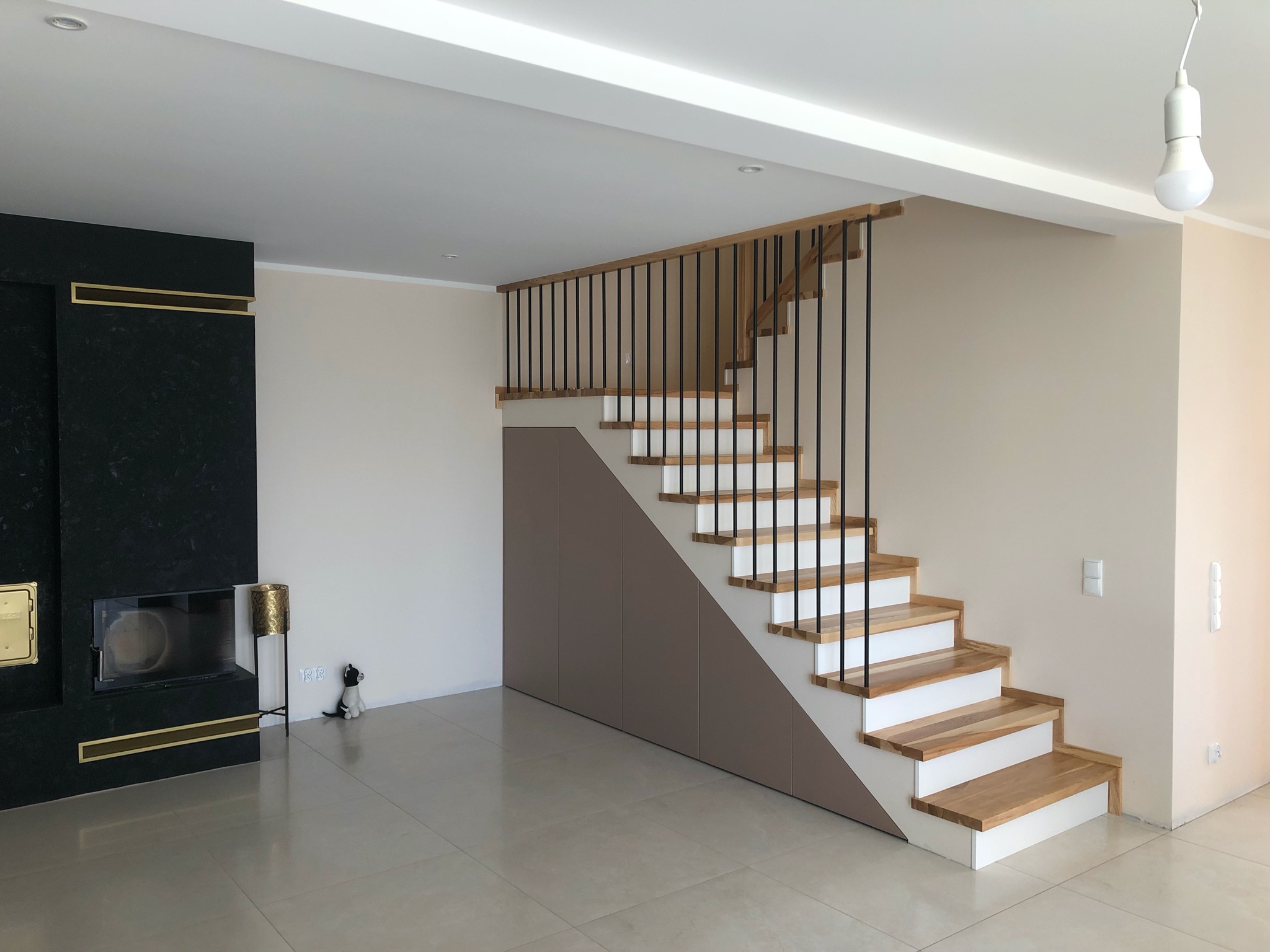 Jasne wnętrze z drewnianymi schodami o białych podstopnicach i czarnych, pionowych balustradach, zintegrowane z zabudową szaf pod schodami, obok czarny kominek z elementami złota i ceramiczna...