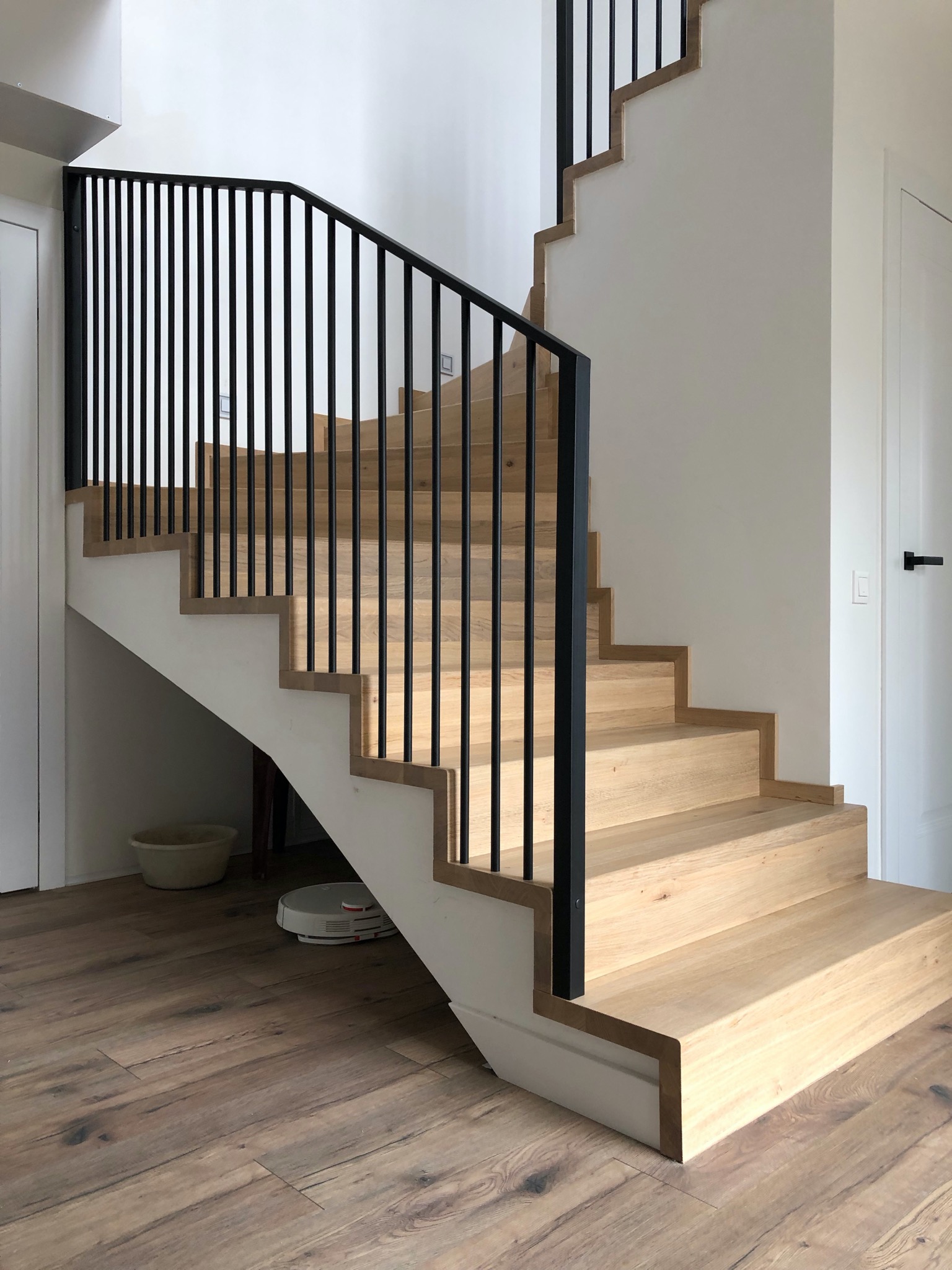 Jasne drewniane schody z czarną metalową balustradą w nowoczesnym wnętrzu, widoczny odkurzacz automatyczny pod schodami.