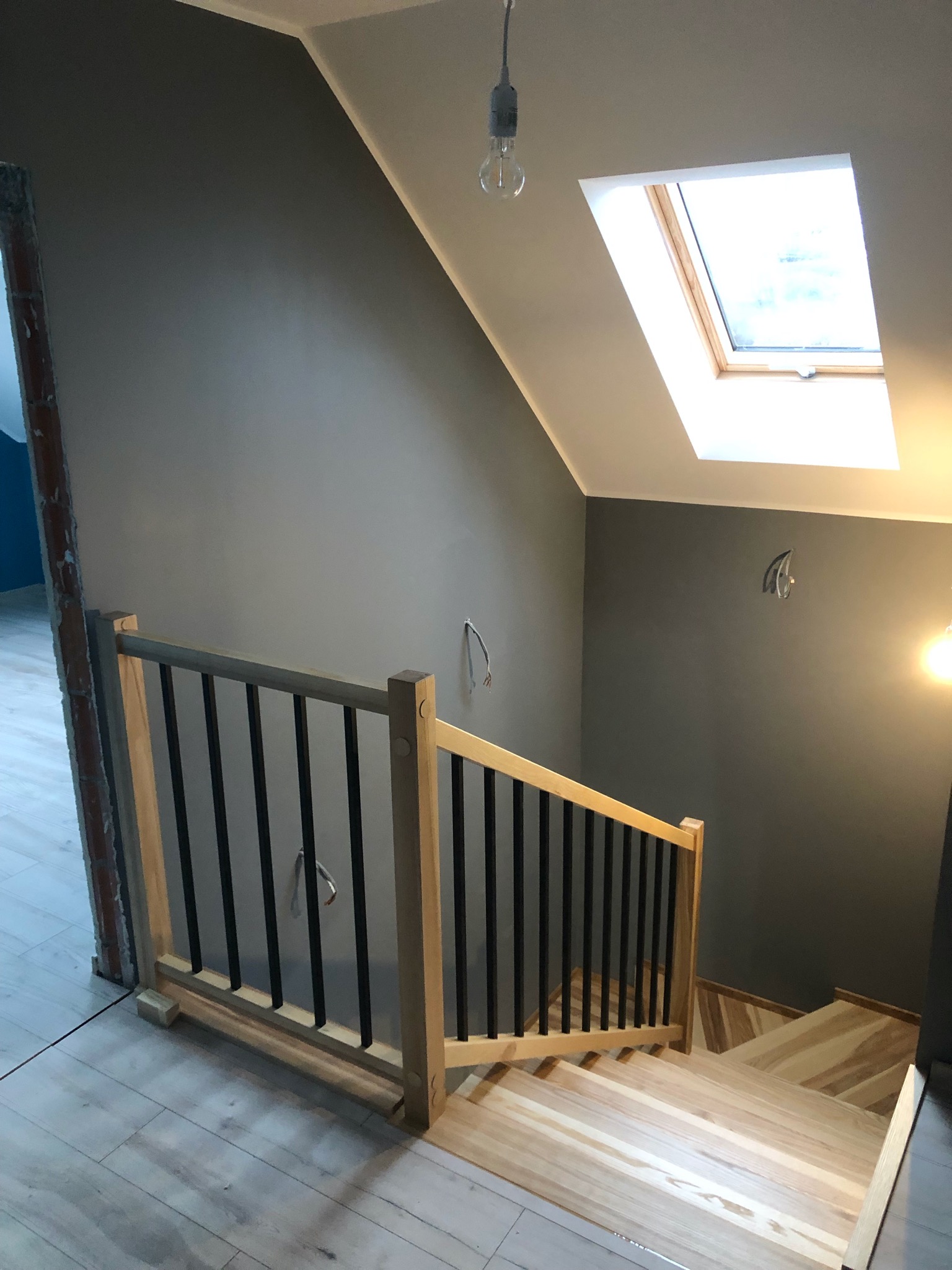 Jasne, drewniane schody z czarnymi metalowymi prętami balustrady w nowoczesnym domu z oknem dachowym i surowym wykończeniem ściany przy schodach.