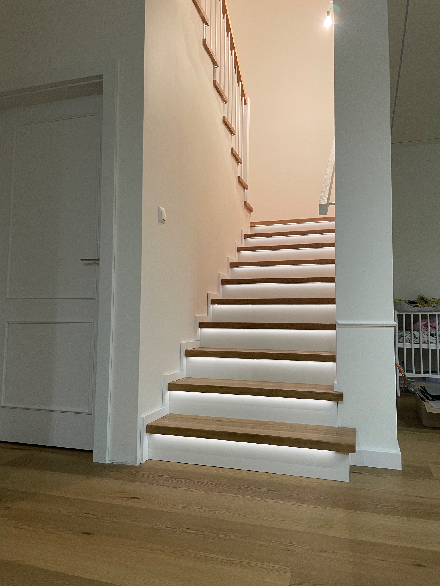 Drewniane schody z białymi podstopnicami i subtelnym oświetleniem LED, prowadzące do góry. Widoczna drewniana podłoga i białe drzwi w tle.