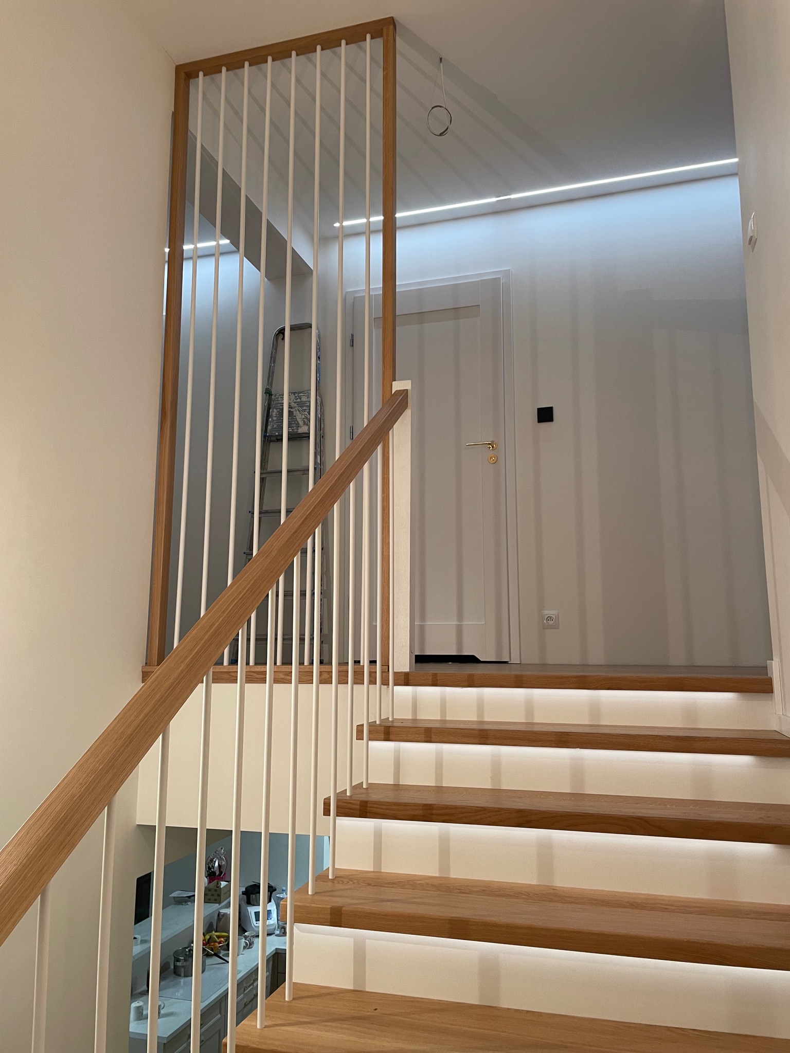 Nowoczesne schody z podświetleniem LED i balustradą z białych prętów w drewnianej ramie. Widoczny fragment poręczy i drzwi w tle. Minimalistyczny design.