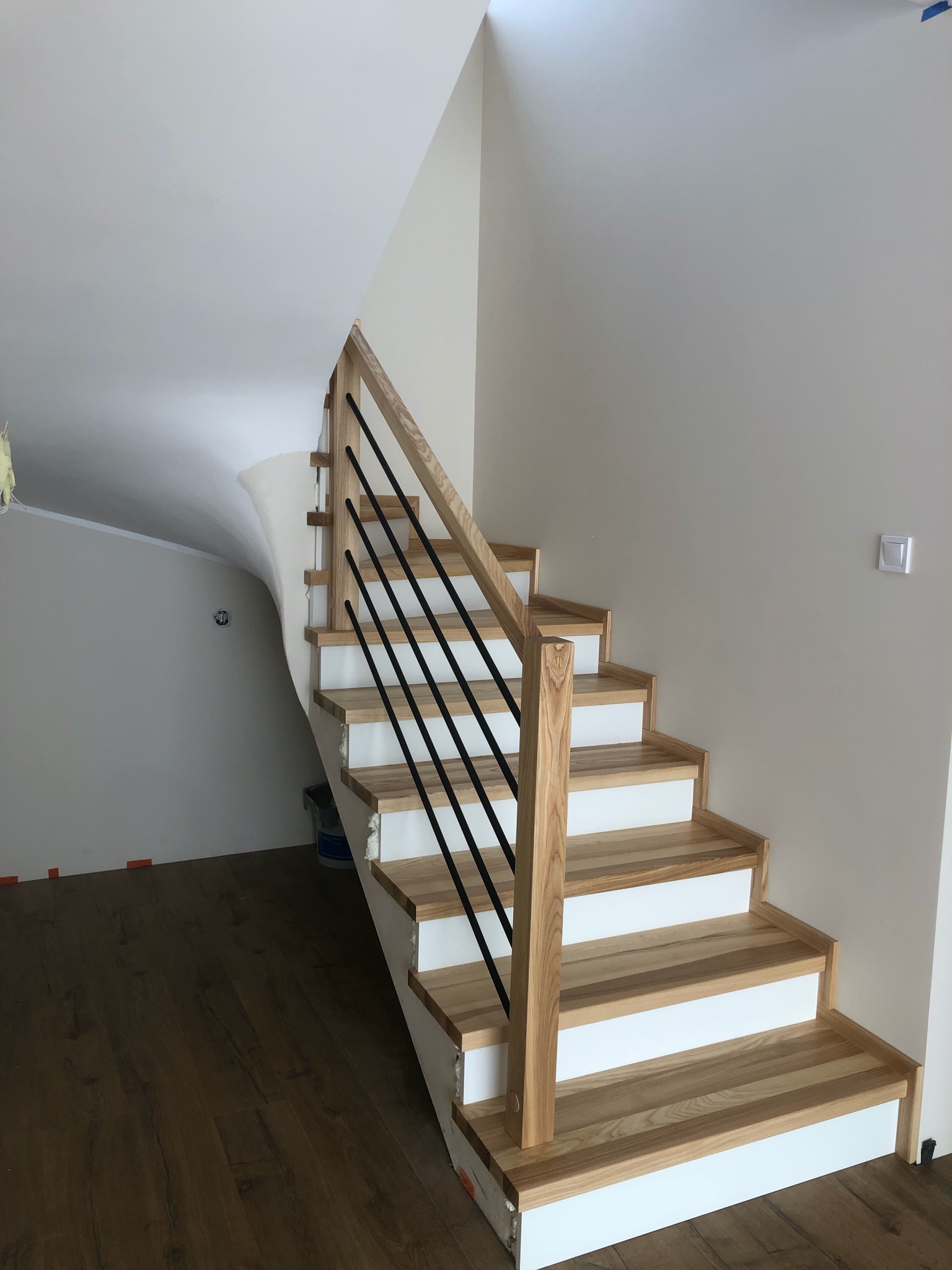 Jasne schody z drewnianymi stopniami i czarną, metalową balustradą w nowoczesnym wnętrzu. Widoczne wykończenia ścian i podłogi.