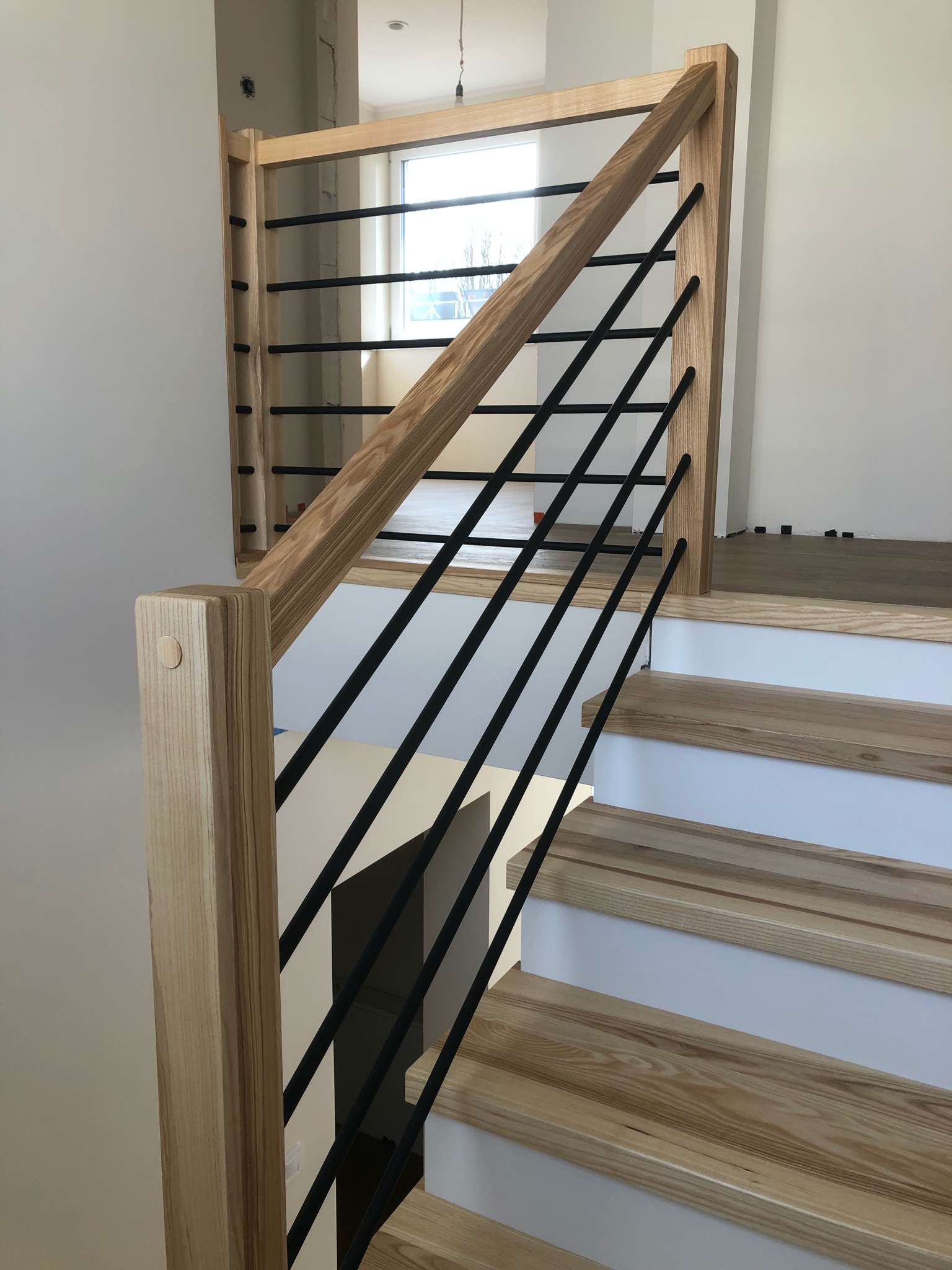 Jasne, drewniane schody z czarnymi, metalowymi prętami balustrady. Widoczne detale wykończenia i naturalna tekstura drewna. Minimalistyczny design w nowoczesnym wnętrzu.