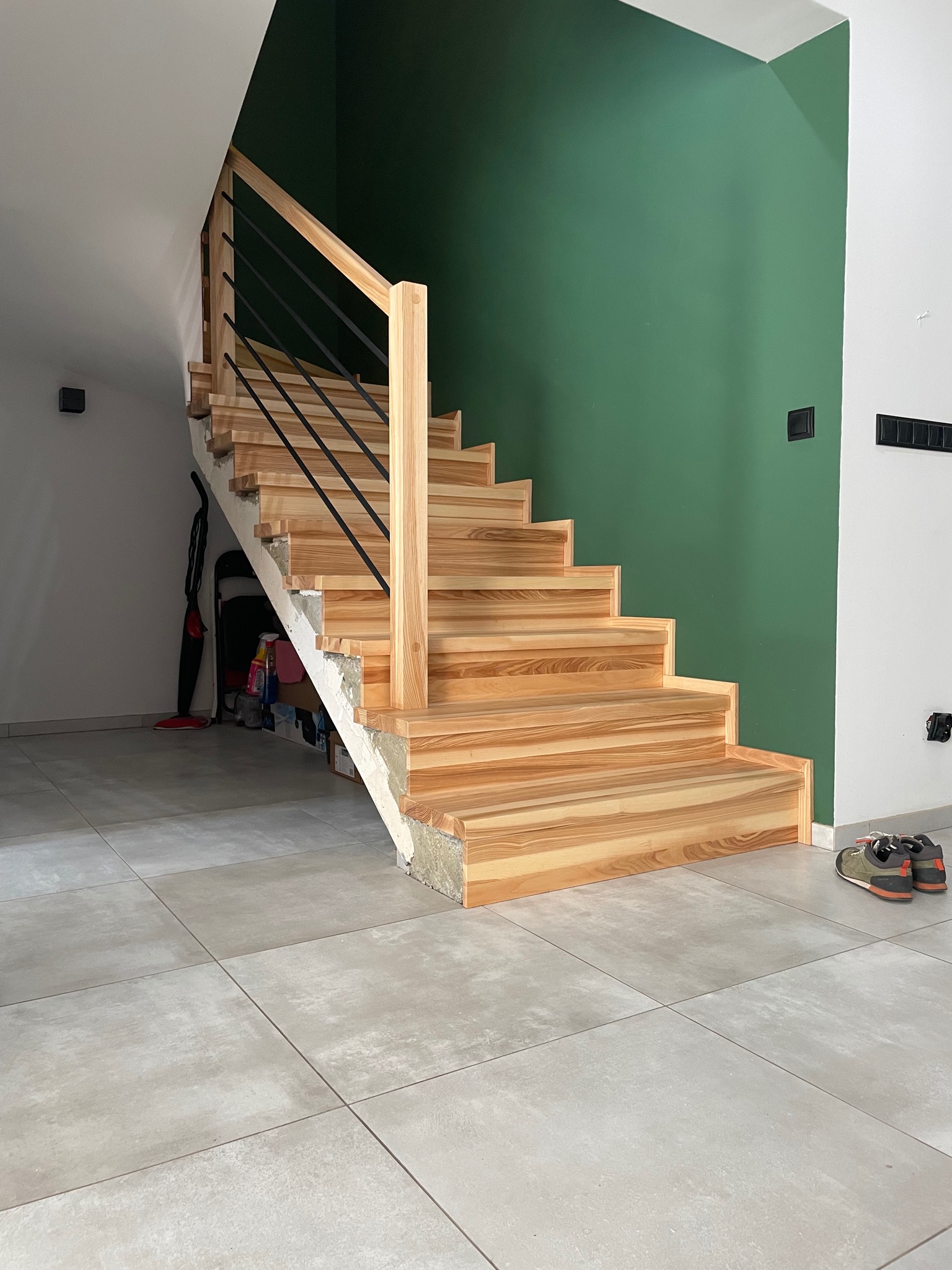 Jasne, drewniane schody z czarnymi, metalowymi prętami balustrady, na tle ściany w kolorze butelkowej zieleni. Widoczne buty na podłodze i przedmioty pod schodami.