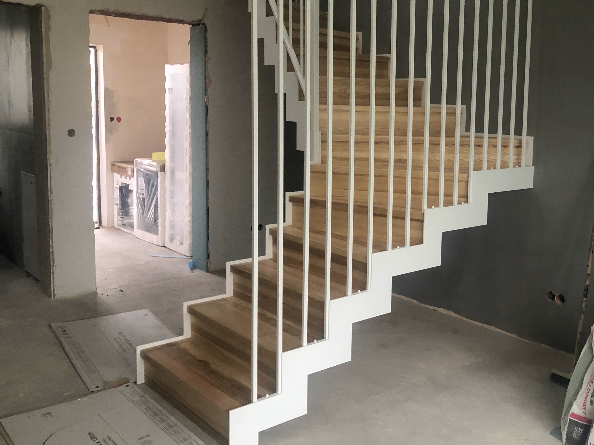 Nowoczesne schody z drewnianymi stopniami i białą, minimalistyczną balustradą w surowym wnętrzu. Widoczne przygotowania do wykończenia pomieszczenia.