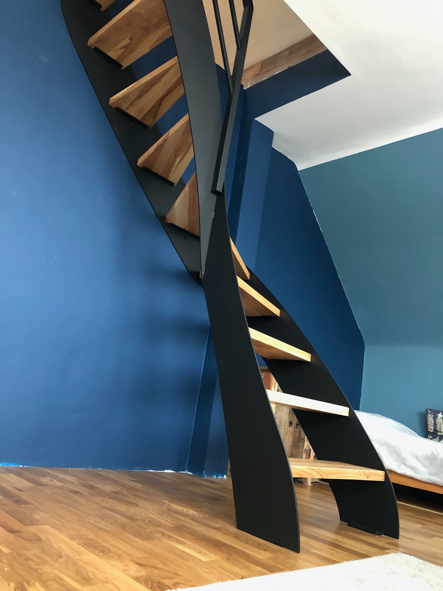 Nowoczesne schody z drewnianymi stopniami i czarną, metalową konstrukcją na tle niebieskiej ściany. Widok z dołu, ukazujący unikalny design i solidne wykonanie.