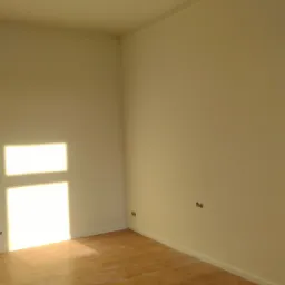 Pusty pokój po malowaniu ścian na biało, z drewnianą podłogą i dekoracyjnym sztukaterią na suficie i pod sufitem, widoczne gniazdka elektryczne.