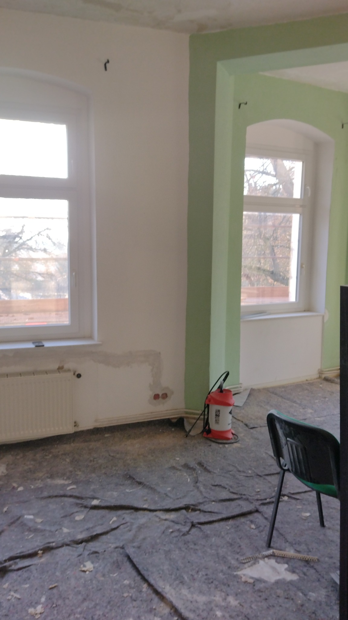 Wnętrze pokoju w trakcie remontu: białe ściany częściowo przygotowane do malowania, okna z białymi ramami, podłoga zabezpieczona folią malarską, odkurzacz przemysłowy w tle.