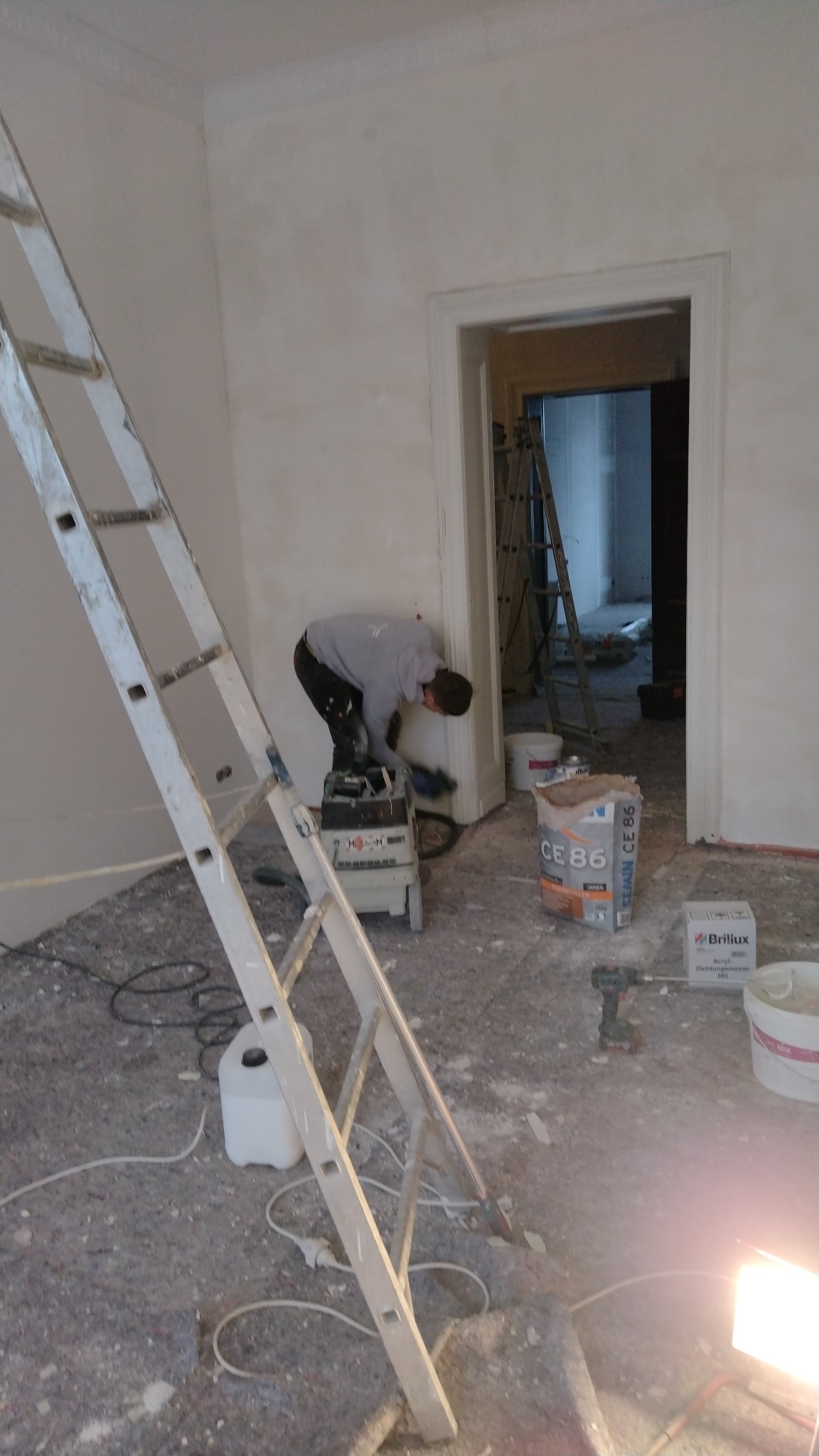 Pracownik szlifuje ścianę wewnątrz pomieszczenia, drabina oparta o ścianę, widoczne narzędzia i materiały budowlane, worek z gipsem CE 86 i wiadro z farbą Brillux.