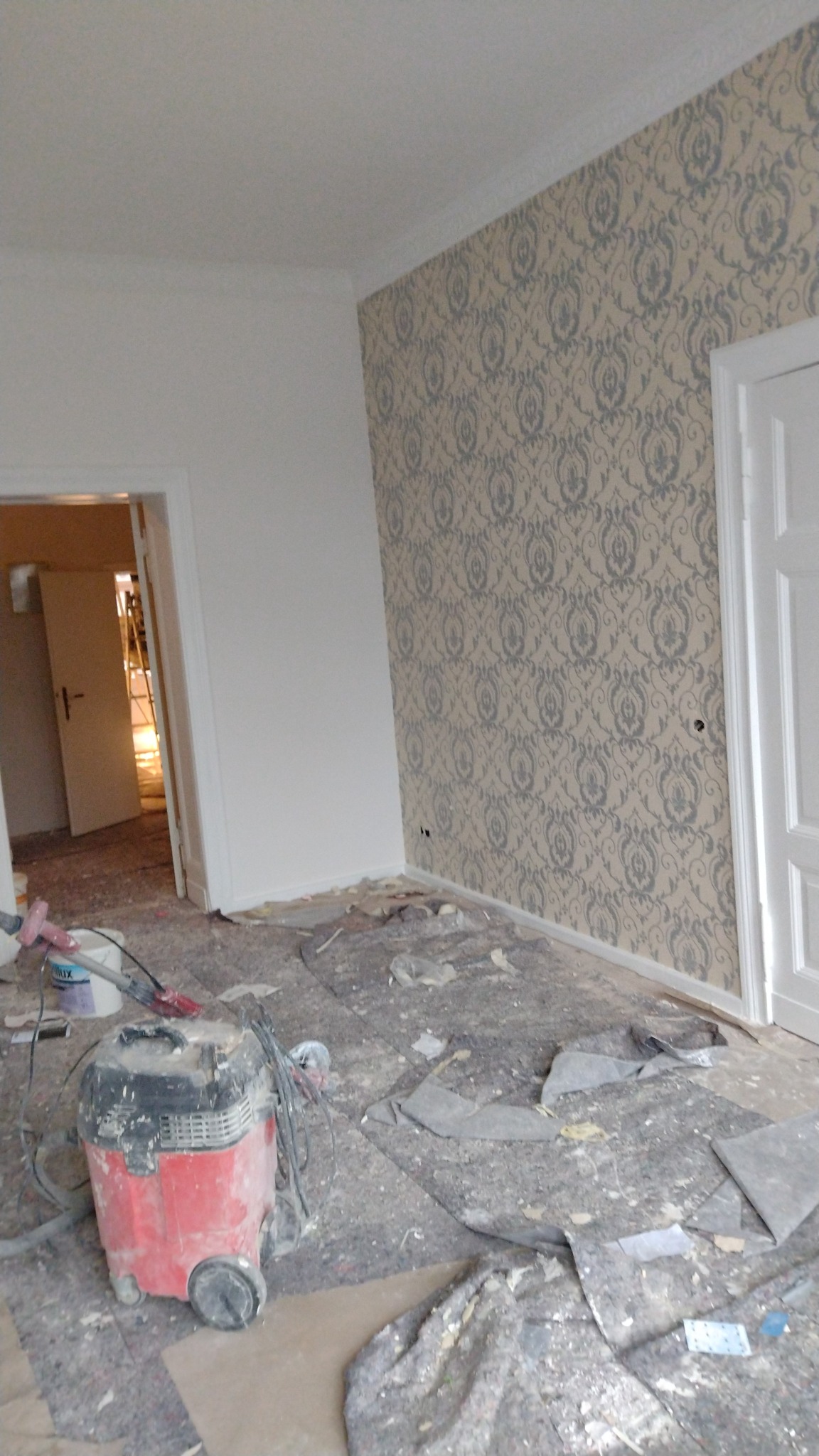 Pokój w trakcie remontu: ściana z wzorzystą tapetą sąsiaduje z świeżo pomalowaną białą ścianą. Na podłodze rozłożona folia ochronna, widoczny odkurzacz przemysłowy i resztki materiałów budowlanych.