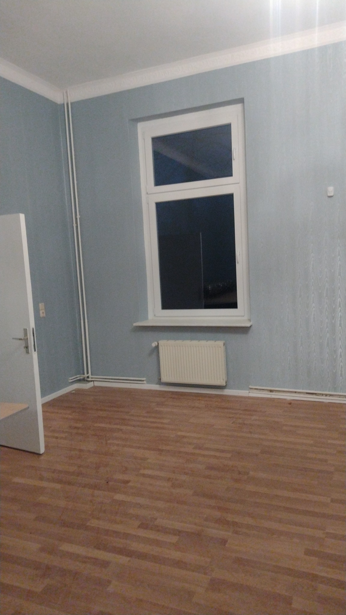 Pusty pokój z niebieskimi ścianami, białym oknem, grzejnikiem i drewnianą podłogą. Widoczny fragment otwartych drzwi.