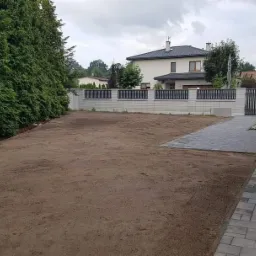 Przygotowany teren pod trawnik, widoczna świeżo wyrównana ziemia, obok ułożona kostka brukowa, w tle żywopłot, ogrodzenie panelowe i dom.