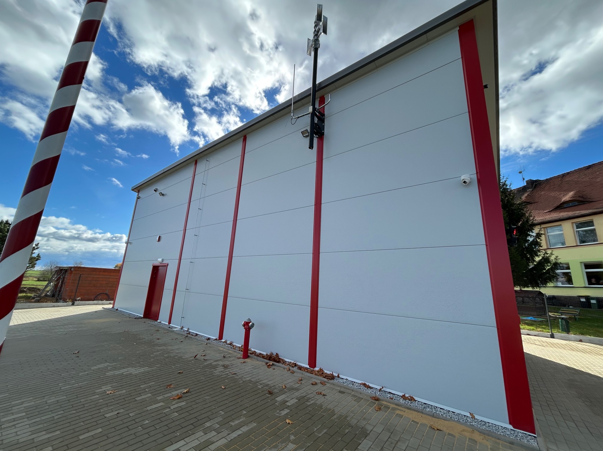 Nowoczesna hala magazynowa z białymi panelami i czerwonymi akcentami, widok z zewnątrz, błękitne niebo z chmurami w tle. Na ścianie antena i kamera.