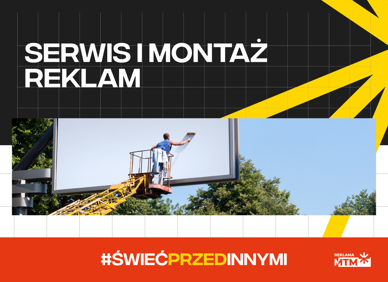 Mężczyzna w podnośniku koszowym czyści duży billboard reklamowy z użyciem wałka, widok z dołu na tle błękitnego nieba i drzew.