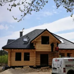 Budowa domu z bali w trakcie, widoczni pracownicy na rusztowaniu montujący elementy dachu, na pierwszym planie samochód dostawczy z logo firmy.