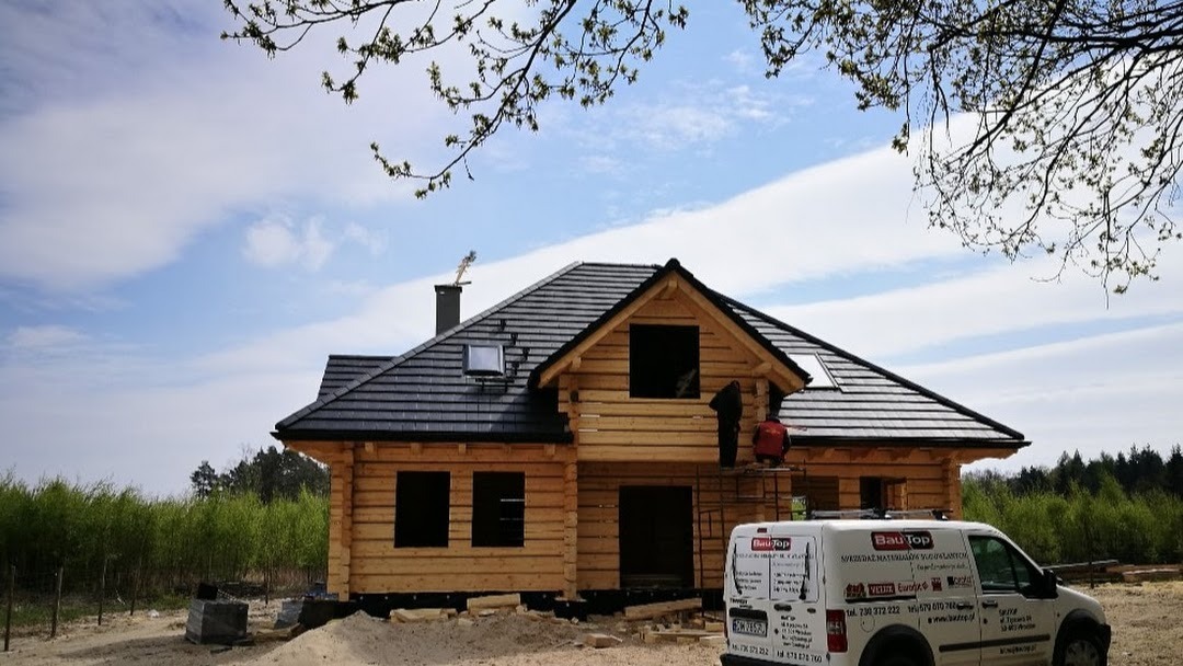 Budowa domu z bali w trakcie, widoczni pracownicy na rusztowaniu montujący elementy dachu, na pierwszym planie samochód dostawczy z logo firmy.