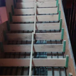 Konstrukcja szalunku schodów żelbetowych z drewnianych desek i prętów zbrojeniowych, widok z góry na surowym etapie budowy.