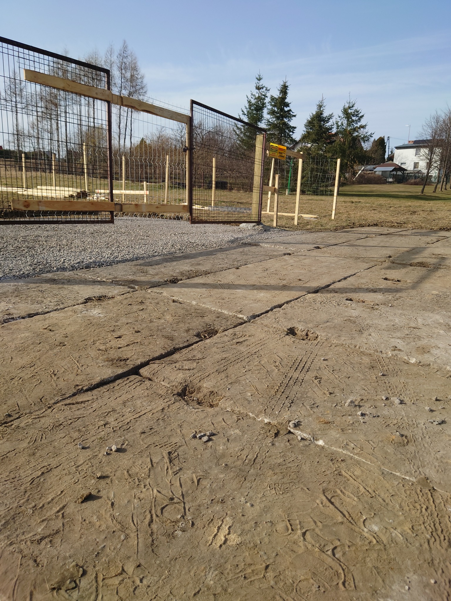 Teren budowy ogrodzenia z metalowej siatki, drewniane elementy konstrukcyjne, betonowe płyty z odciskami opon i butów, w tle drzewa i budynek.