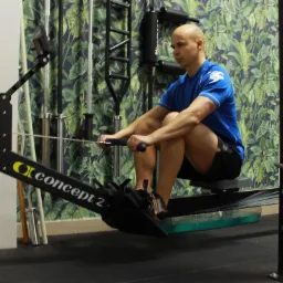 Mężczyzna ćwiczy na ergometrze wioślarskim Concept2 w siłowni z zieloną tapetą w liście.