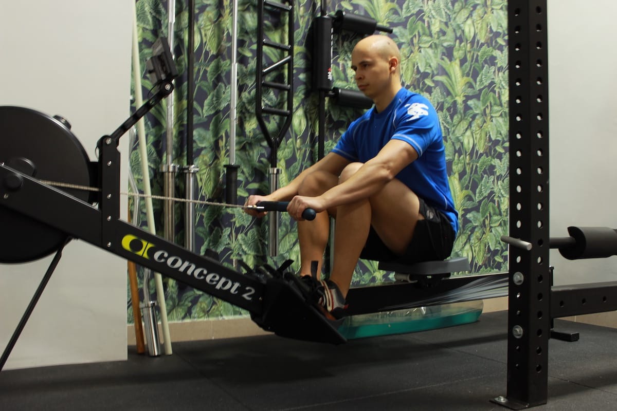 Mężczyzna ćwiczy na ergometrze wioślarskim Concept2 w siłowni z zieloną tapetą w liście.
