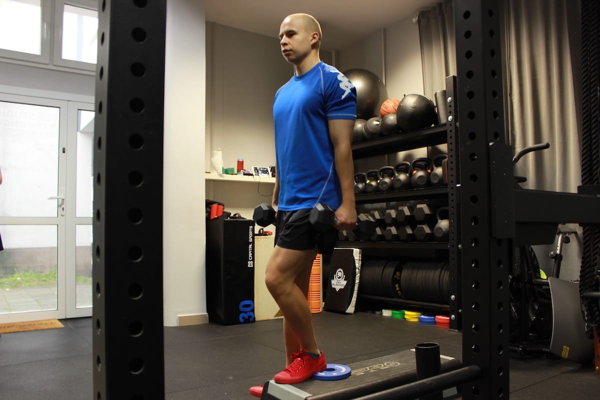 Mężczyzna w niebieskiej koszulce i czerwonych butach wykonuje ćwiczenie z hantlami, stojąc na platformie w siłowni z wyposażeniem w tle.