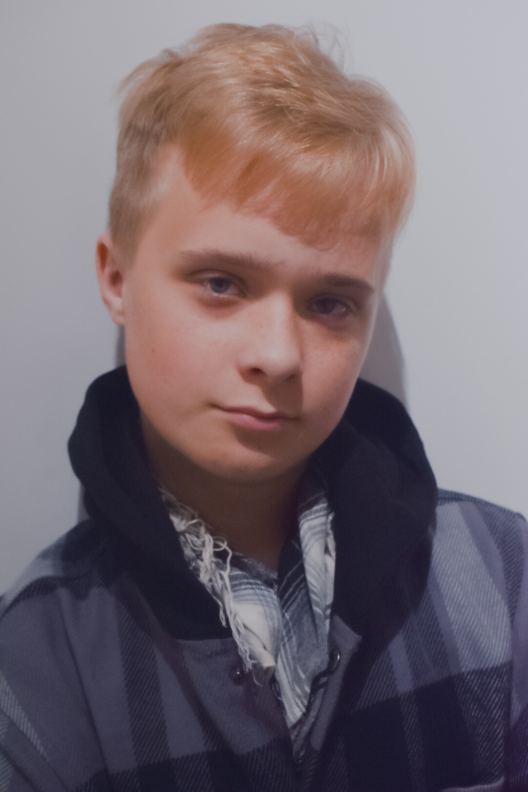 Portret młodego mężczyzny o jasnych włosach, ubrany w szarą koszulę w kratę i czarną bluzę z kapturem, z szalikiem na szyi, na tle jasnej ściany. Umiarkowane oświetlenie.