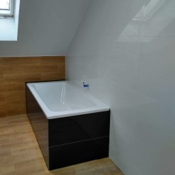 Róża Kołtun - Biała wanna z czarną obudową w łazience na poddaszu z drewnopodobnymi płytkami na podłodze i ścianie, białe płytki na ścianie pod skosem, małe okno dachowe.