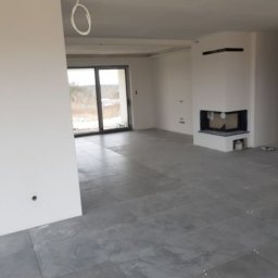 Róża Kołtun - Surowe wnętrze domu w stanie deweloperskim z szarymi płytkami na podłodze, białymi ścianami, kominkiem i widokiem na zimowy krajobraz za oknem balkonowym.