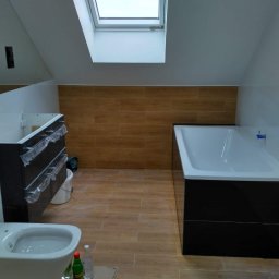 Róża Kołtun - Łazienka na poddaszu w trakcie remontu: widoczna wanna obudowana czarnymi płytkami, bidet z butelkami na podłodze, częściowo zamontowana szafka pod umywalkę z folią ochronną, oraz okno dachowe.
