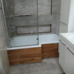 Nowoczesna łazienka z wanną zabudowaną drewnopodobnymi panelami, szklaną kabiną prysznicową i białą umywalką z szafką, wykończona szarą płytką imitującą cegłę.