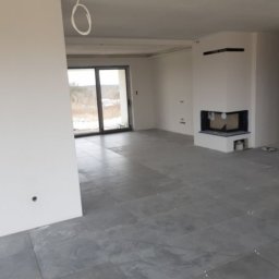 Róża Kołtun - Surowe wnętrze domu w trakcie wykańczania: szare płytki podłogowe z krzyżykami dystansowymi, białe ściany z wystającym kablem, kominek w narożniku i duże okno balkonowe.