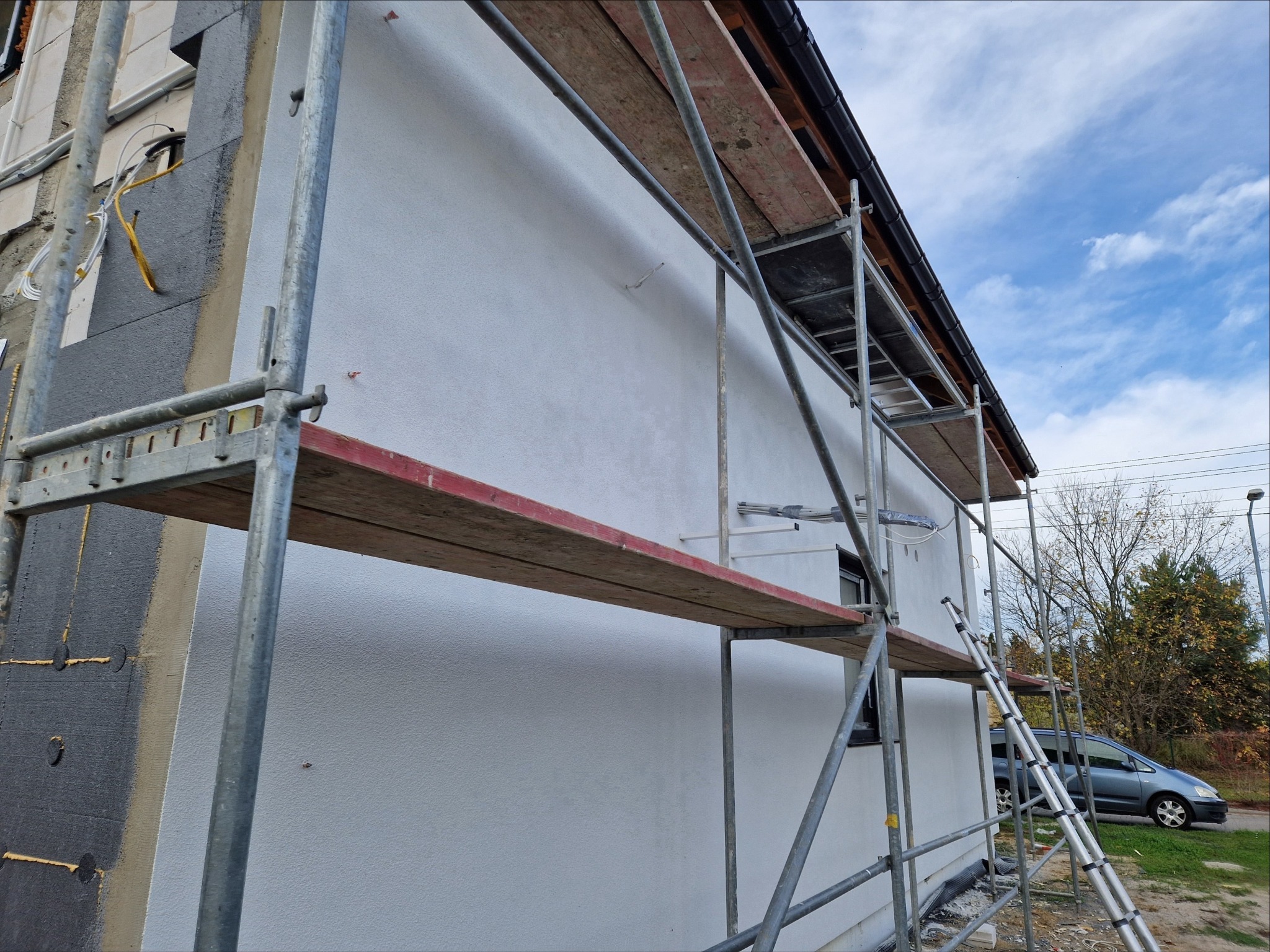 Dom w trakcie ocieplania, widoczne rusztowanie, fragment ściany z szarym ociepleniem i fragment z nałożonym białym tynkiem strukturalnym, błękitne niebo w tle.
