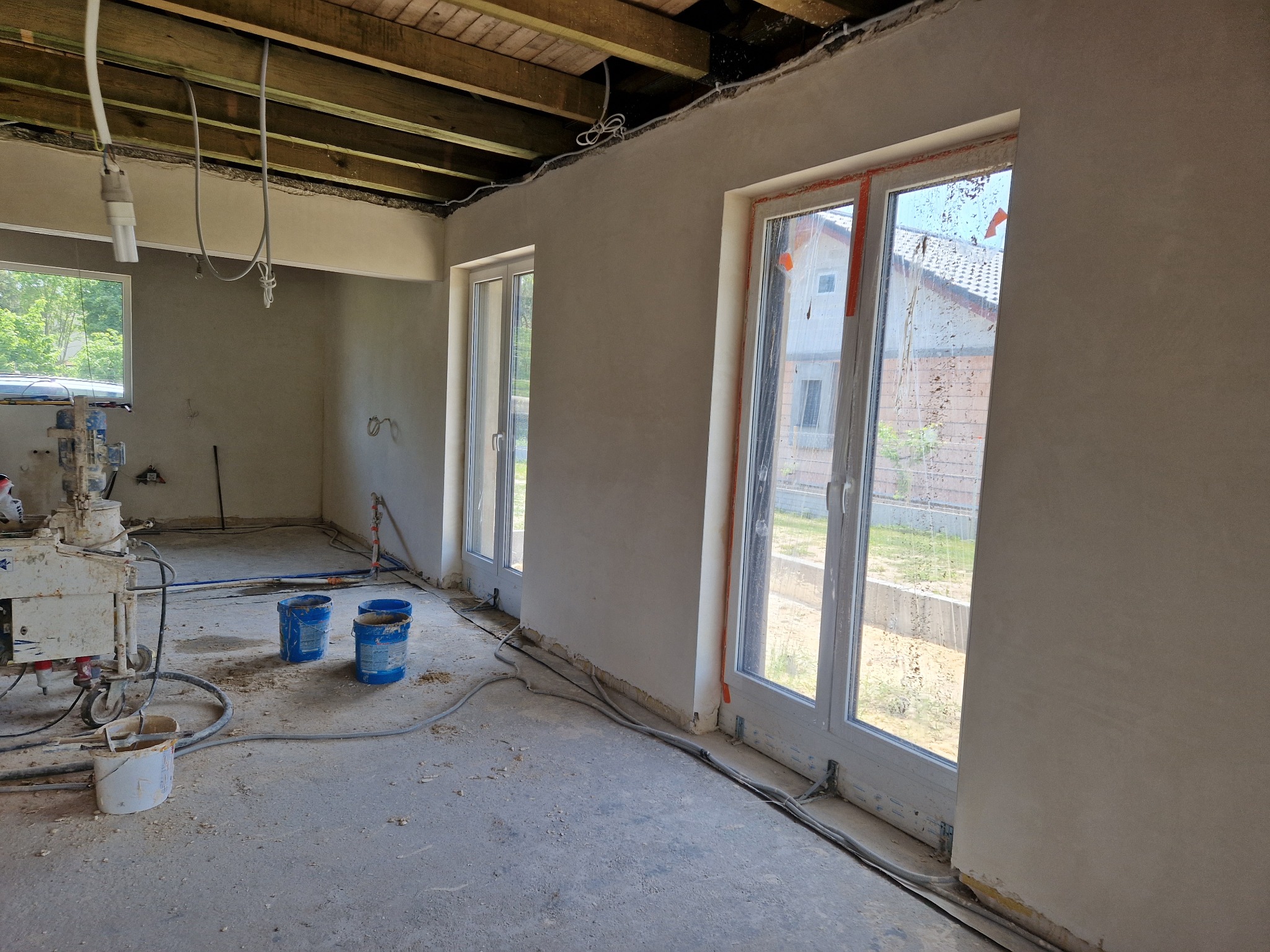 Wnętrze w trakcie prac wykończeniowych: świeżo nałożona szpachla na ścianach, widoczne okna z folią ochronną, maszyna do szpachlowania i niebieskie wiadra na podłodze.