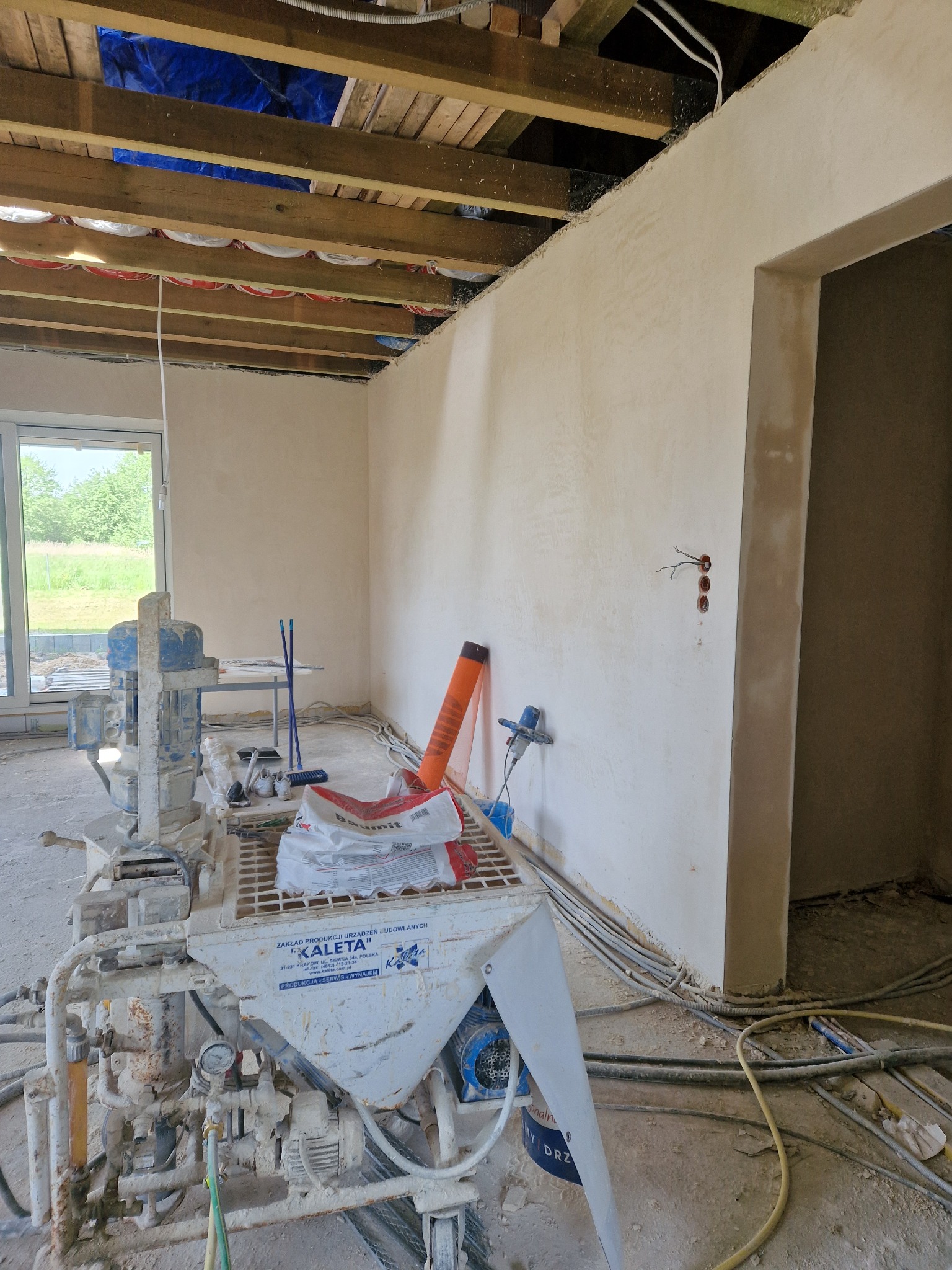 Wnętrze w trakcie prac, ściany po szpachlowaniu, widoczna maszyna do nakładania gładzi marki Kaleta, instalacja elektryczna wystająca ze ściany, surowy strop z drewnianych belek.