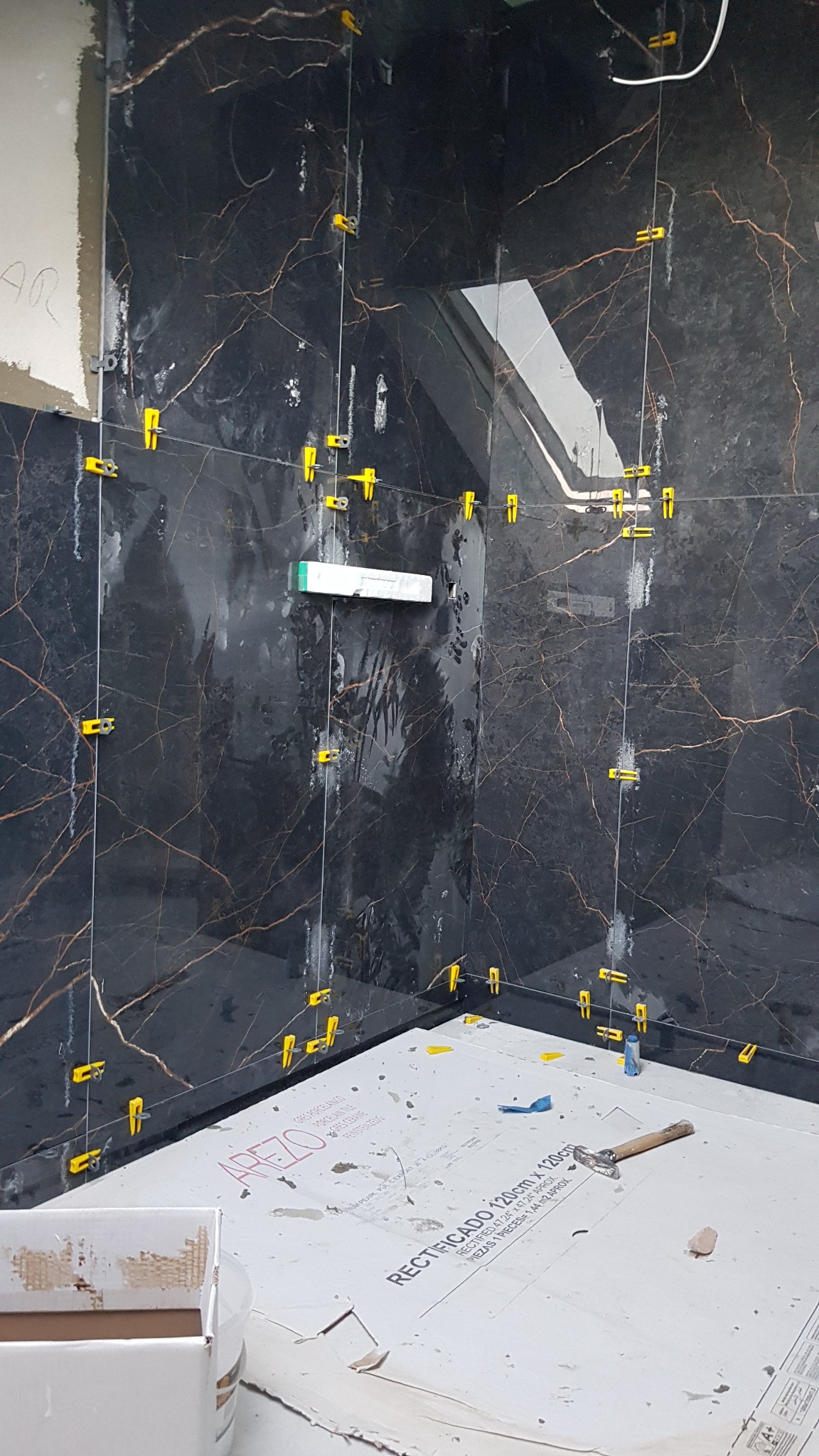 Montaż płytek imitujących czarny marmur z żółtymi krzyżykami dystansowymi w rogu łazienki, widoczne narzędzia i opakowania płytek.