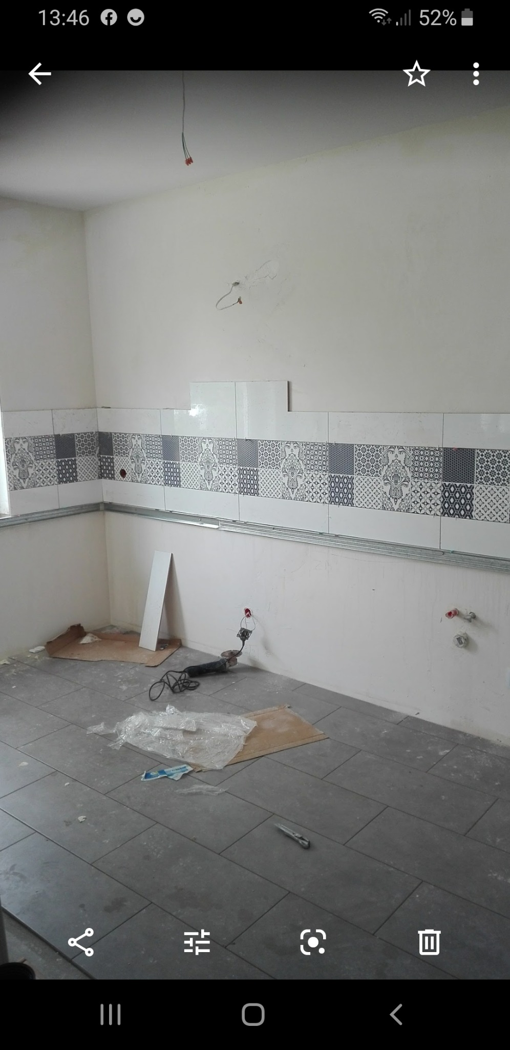 Wnętrze łazienki w trakcie remontu: częściowo położone płytki wzorzyste na ścianie, szare płytki podłogowe, widoczne narzędzia i materiały budowlane, niedokończone instalacje.