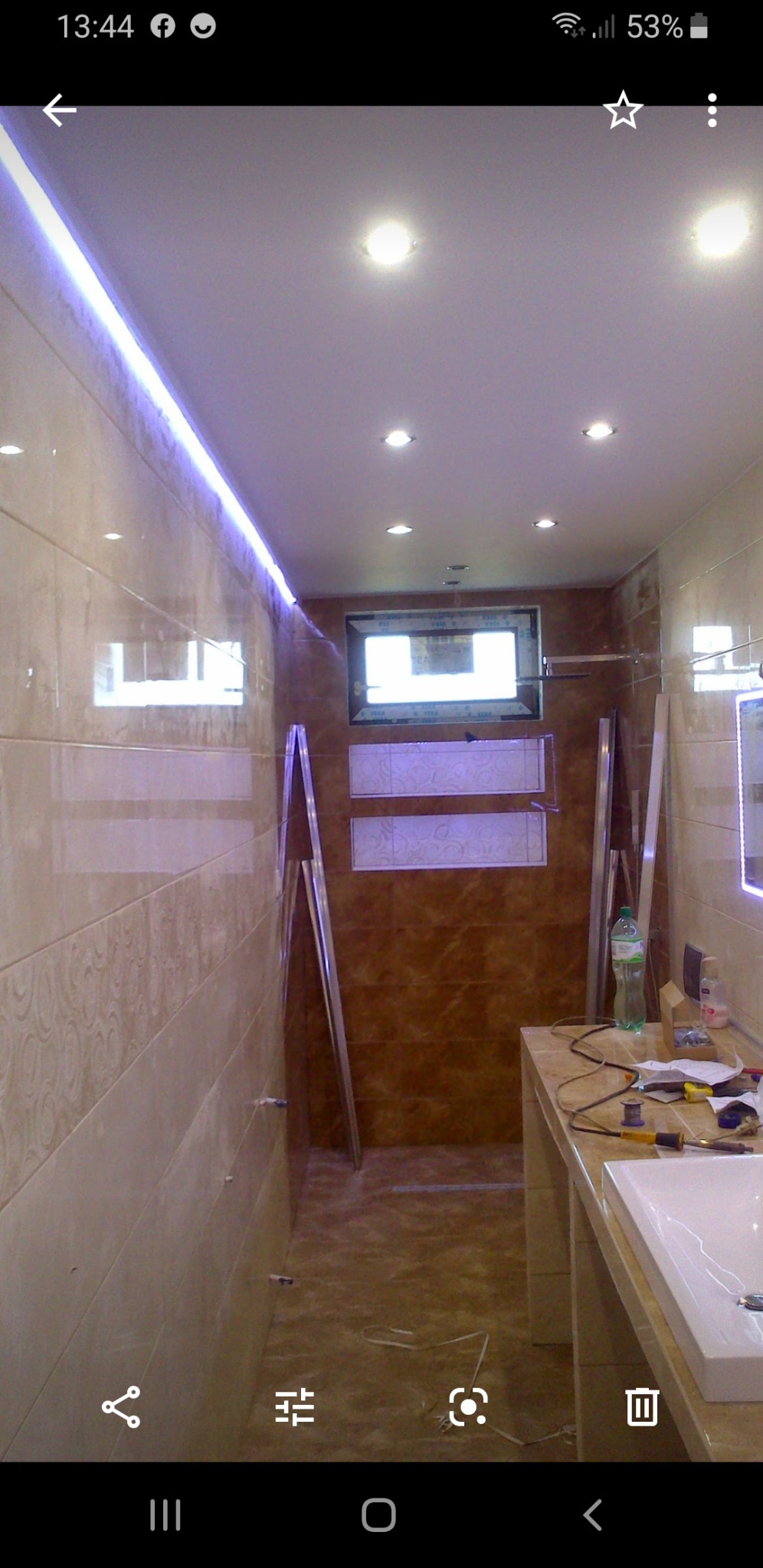 Łazienka w trakcie remontu, widoczne płytki na ścianach i podłodze, podświetlenie LED przy suficie, niedokończona zabudowa wnęki, lustro i umywalka, narzędzia na blacie.