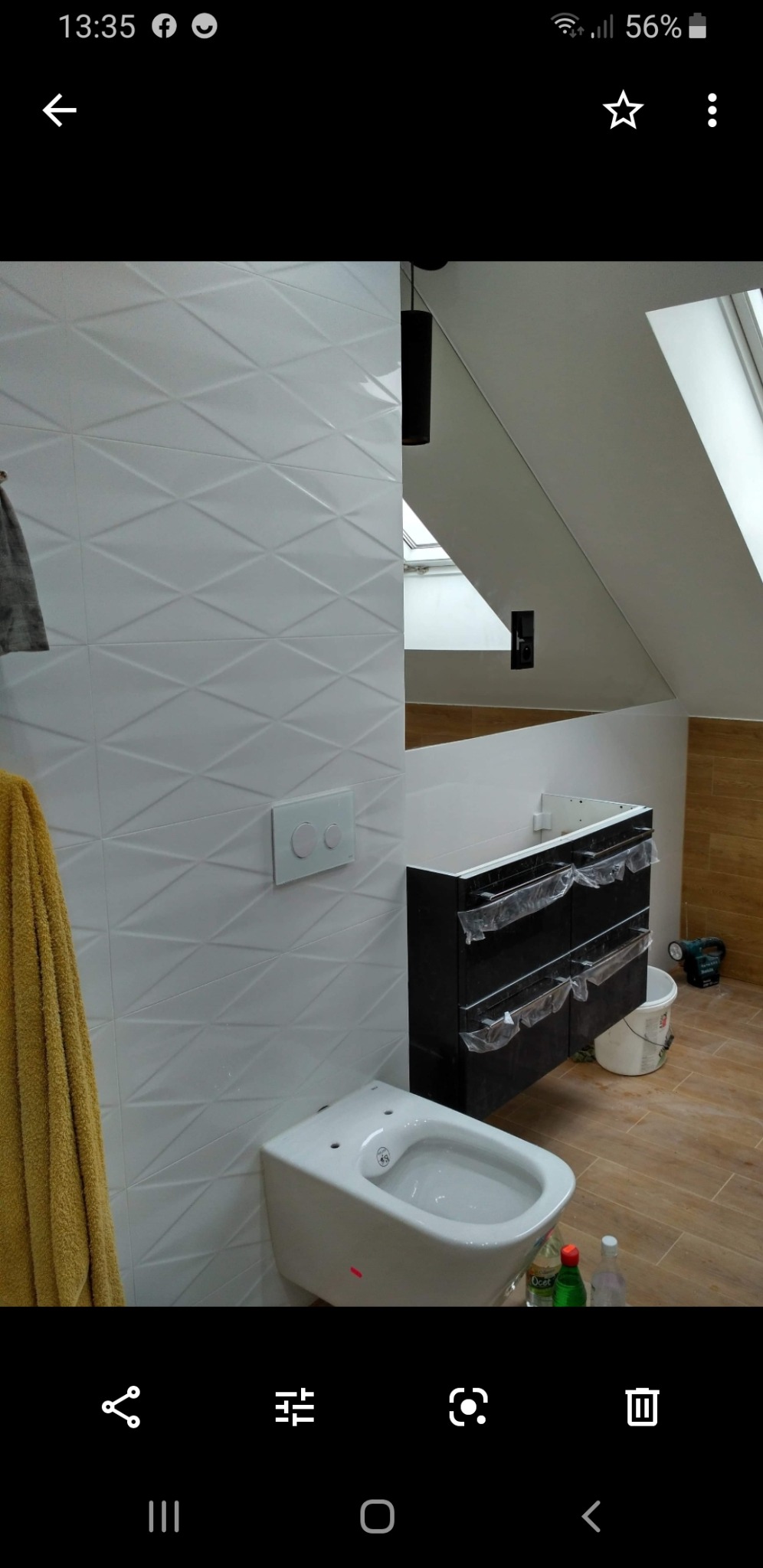 Nowoczesna łazienka w trakcie remontu: biała, trójwymiarowa glazura na ścianie, podwieszana toaleta bez deski, czarna szafka pod umywalkę zabezpieczona folią, drewnopodobne panele na podłodze, okno...