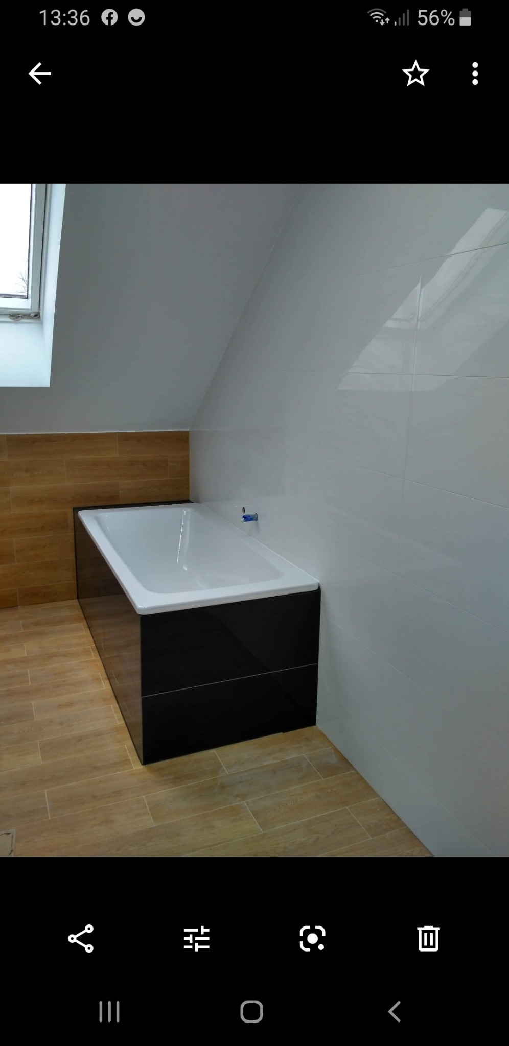 Biała wanna z czarną obudową w łazience na poddaszu z drewnopodobnymi płytkami na podłodze i ścianie, białe płytki na ścianie pod skosem, małe okno dachowe.