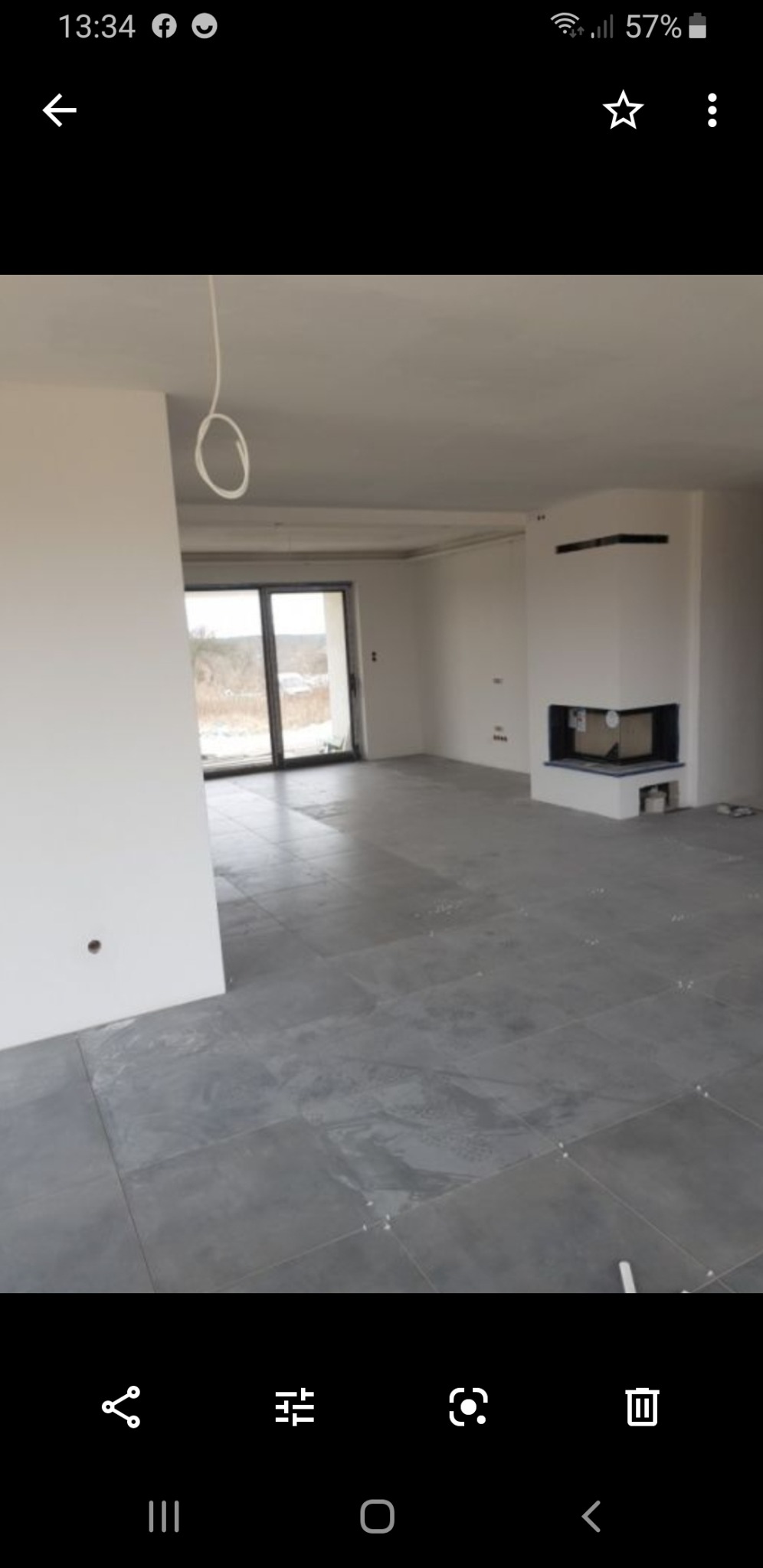 Surowe wnętrze domu w stanie deweloperskim z szarymi płytkami na podłodze, białymi ścianami, kominkiem i widokiem na zimowy krajobraz za oknem balkonowym.