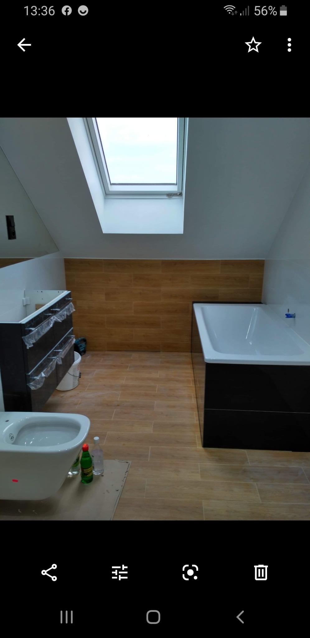 Łazienka na poddaszu w trakcie remontu: widoczna wanna obudowana czarnymi płytkami, bidet z butelkami na podłodze, częściowo zamontowana szafka pod umywalkę z folią ochronną, oraz okno dachowe.