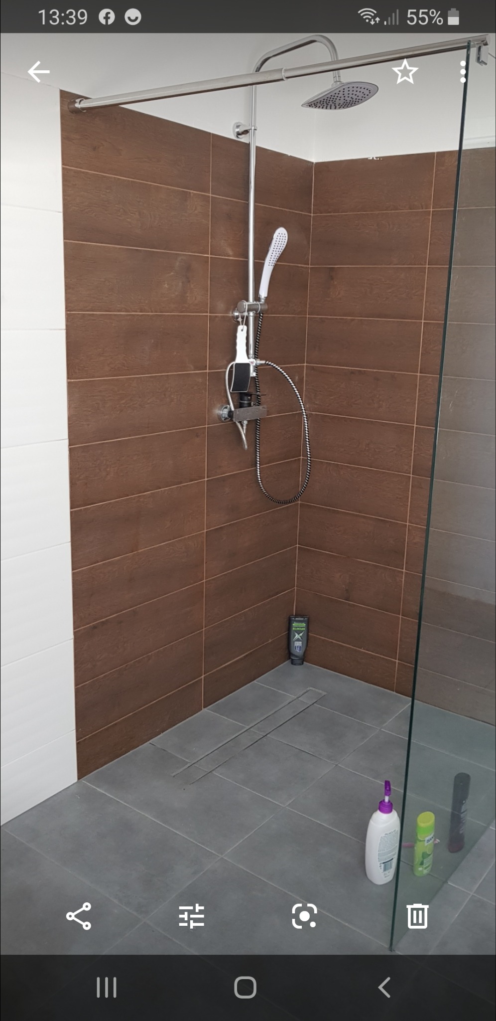 Nowoczesna kabina prysznicowa z brązowymi płytkami imitującymi drewno, szara podłoga z odpływem liniowym, oraz szklana ściana. Widoczne kosmetyki w rogu i przy szklanej ścianie.