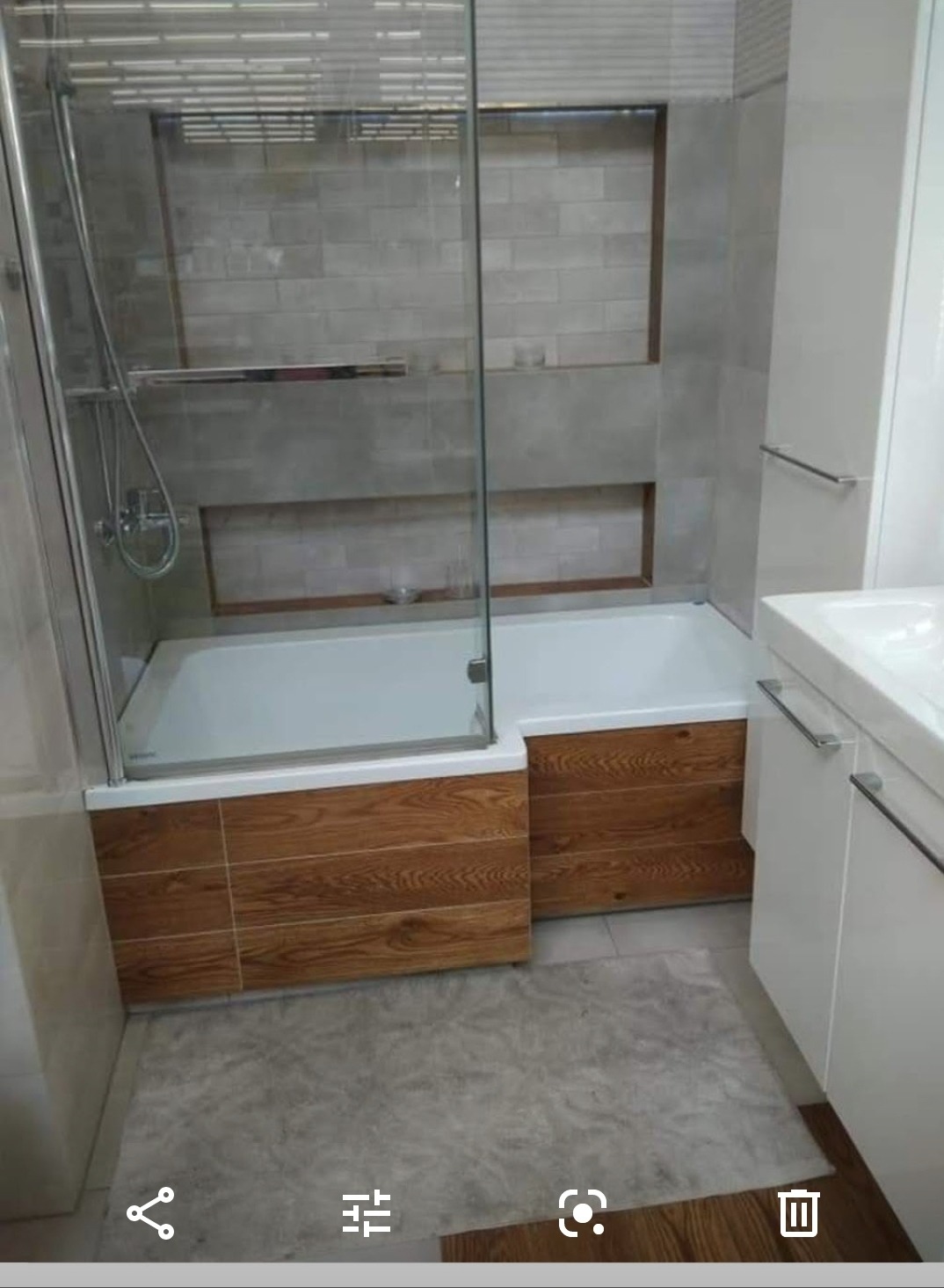 Nowoczesna łazienka z wanną zabudowaną drewnopodobnymi panelami, szklaną kabiną prysznicową i białą umywalką z szafką, wykończona szarą płytką imitującą cegłę.