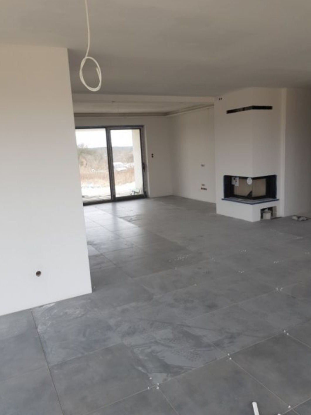 Surowe wnętrze domu w trakcie wykańczania: szare płytki podłogowe z krzyżykami dystansowymi, białe ściany z wystającym kablem, kominek w narożniku i duże okno balkonowe.