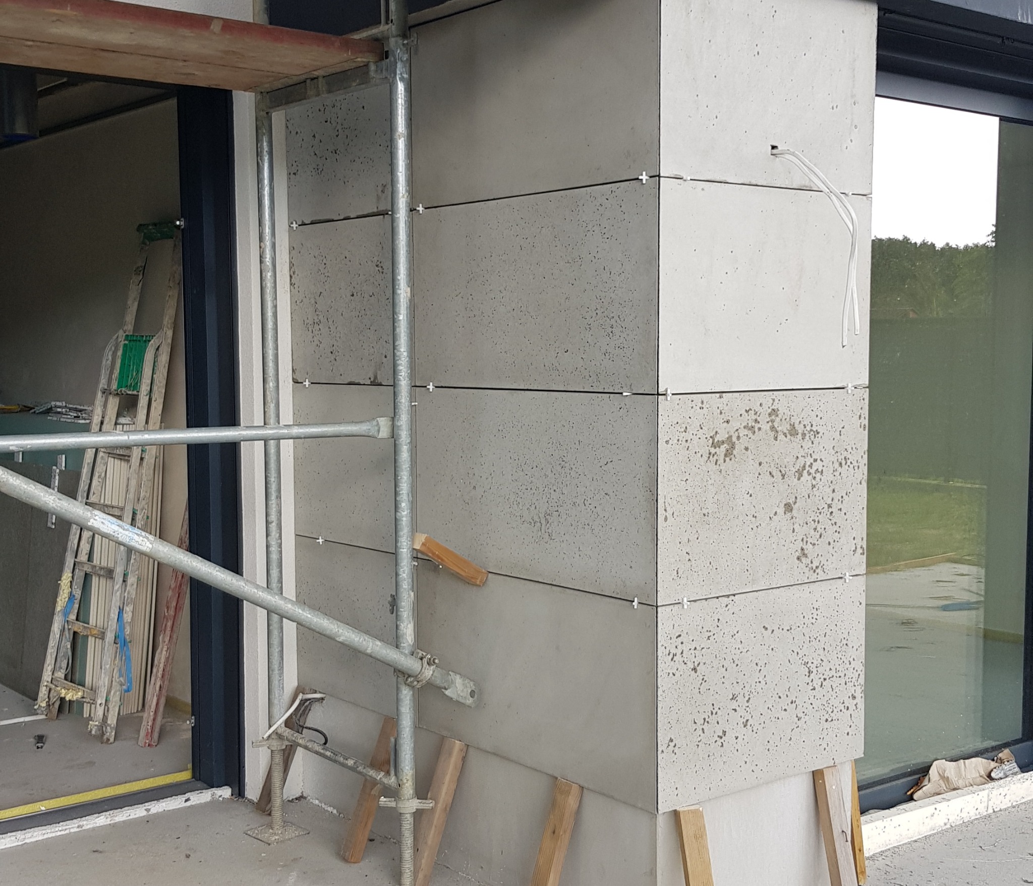 Montaż elewacyjnych płyt betonowych na rogu budynku, widoczne rusztowanie, poziomica klinowa i przewody elektryczne wystające ze ściany.