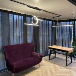 Faanbud - Nowoczesne wnętrze biurowe z bordową sofą, drewnianym biurkiem z czarnymi nogami, panelami podłogowymi w jodełkę i minimalistycznymi lampami wiszącymi na tle zasłon.