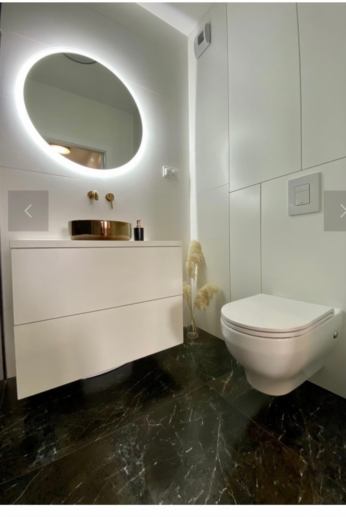 Nowoczesna łazienka z czarną marmurową podłogą, białą szafką pod umywalką z misą w kolorze złota i podświetlanym owalnym lustrem, minimalistyczna toaleta wisząca.
