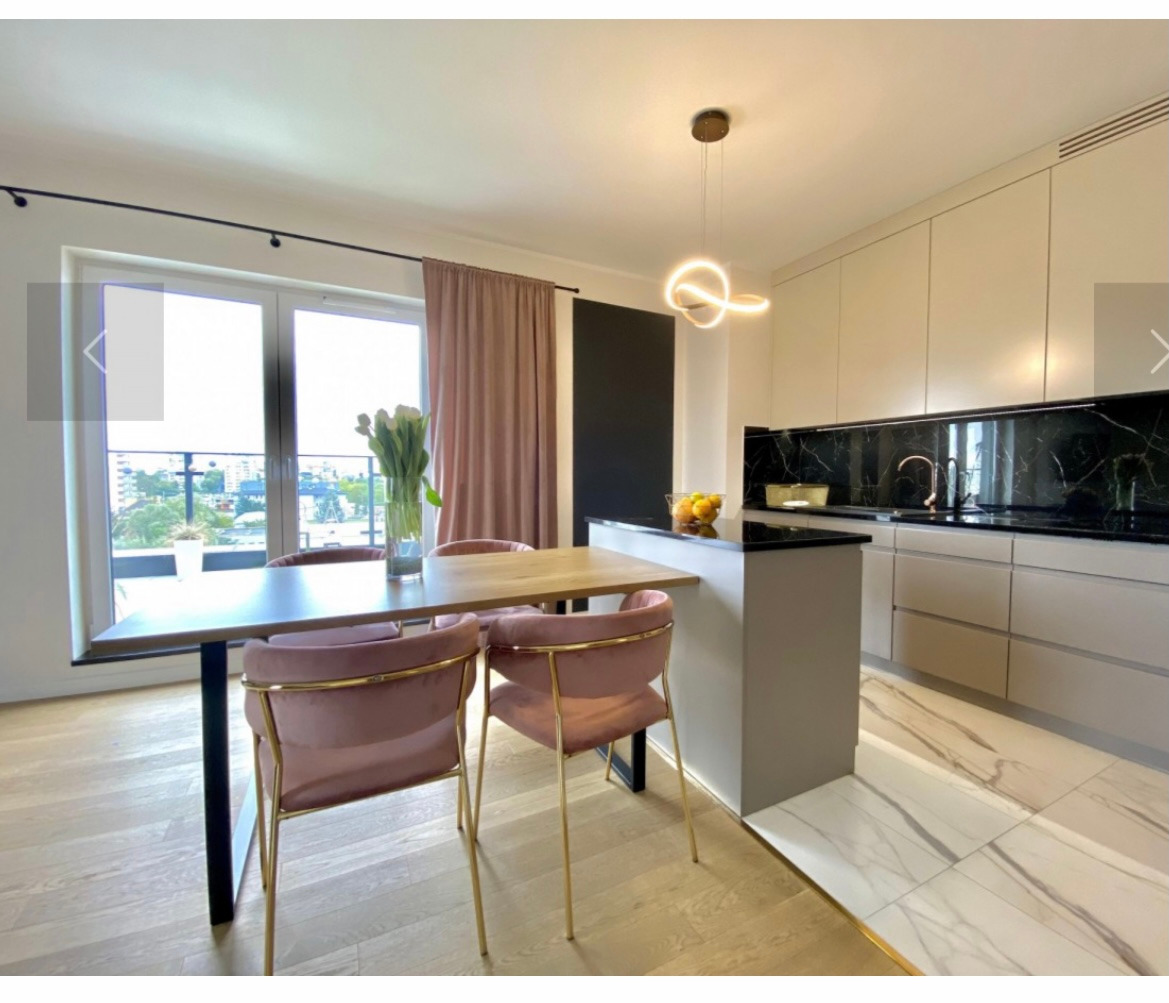 Jasna kuchnia z wyspą, drewnianym blatem stołu i różowymi krzesłami na złotych nogach, oświetlona nowoczesną lampą wiszącą.